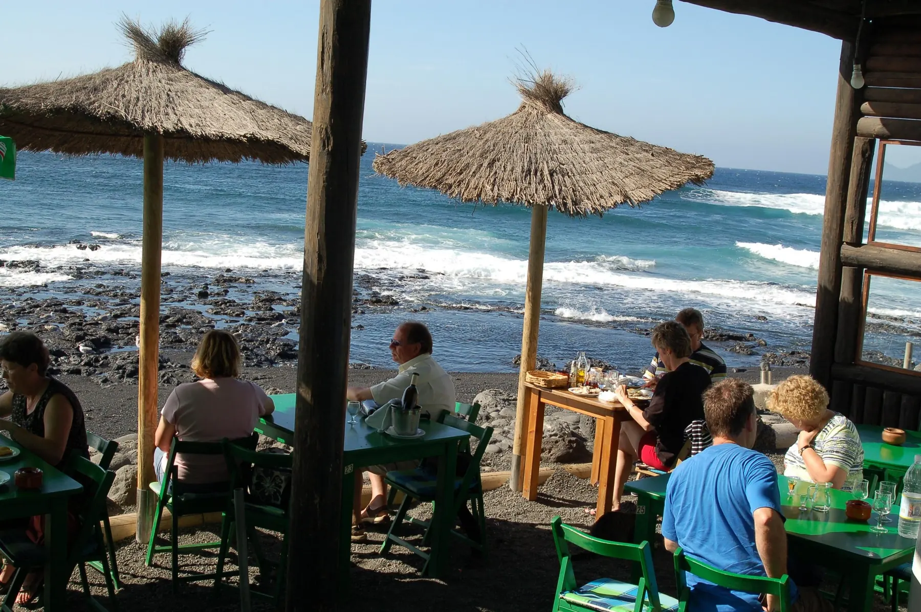 Terraza del Restaurante Bogavante con mesas verdes y sombrillas de paja sobre la arena negra, frente a las olas de El Golfo